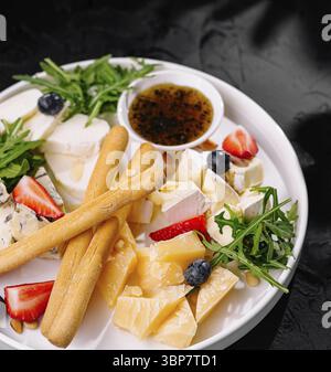 Food, Elegant assortment of fine cheeses paired with fresh fruits, breadsticks, and arugula on a white plate against a dark background Stock Photo