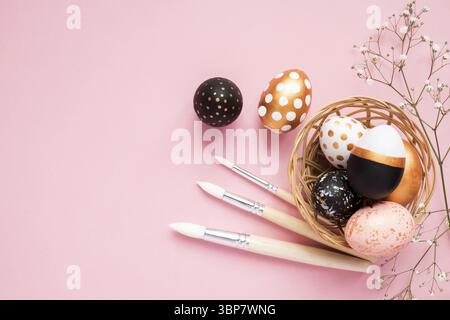 Top view of wooden painted eggs in gold, black and rose colors with branch of gypsophila and paint brushes on pink background. Happy easter background Stock Photo