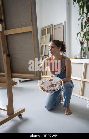 Calm female artist sits on knee holds palette brush works out small details on huge painting creates piece of art. Focused woman listening to music in Stock Photo