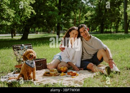 Young Diverse Couple Sitting Together Having Picnic with Dog in Park Stock Photo