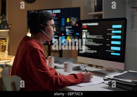 Biracial man wearing headset sitting at desk operating computer with scam call center interface visible on monitor holding pen and paper Stock Photo