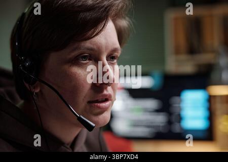 Portrait of Caucasian woman wearing headset working at computer in scam call center, blurred monitor with data visible in background Stock Photo