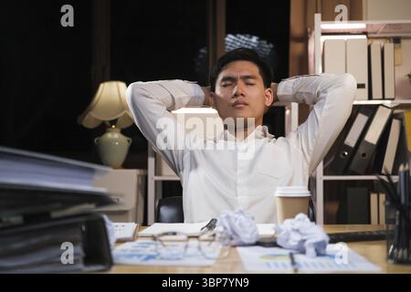 Asian man working late sitting on desk in office overtime at night. Business man feeling tired and stress for overload job Stock Photo