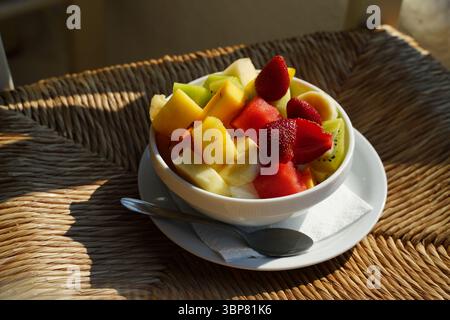 Freshly prepared fruit salad featuring vibrant strawberries, kiwi, pineapple, and watermelon in a white bowl on a wicker surface. A silver spoon rests Stock Photo