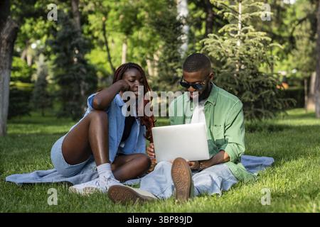 Busy black couple, male and female, engaged with laptop, viewing movies and browsing web, exchanging ideas in park on summer picnic. African students Stock Photo