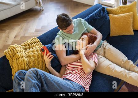 Caring man gentle hug wife enjoying leisure time together, resting on sofa. Smiling woman lying on loving husband laps at home. Romantic relationship. Stock Photo