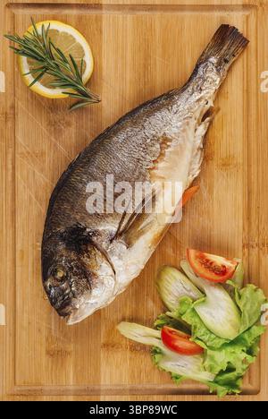 Food, Grilled dorado fish lying on wooden cutting board with vegetables and lemon Stock Photo