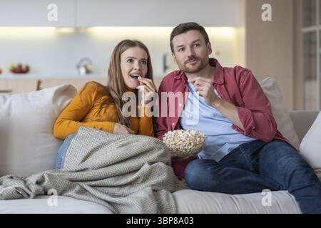 Intrigued couple sitting on sofa, watching favourite humorous show, comedy on TV, eating popcorn, relaxing during weekend. Spending free time, pastime Stock Photo