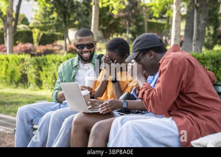 Overjoyed african american girls and guys students sitting in park with laptop laughing together watching videos. Happy four diversity black friends h Stock Photo