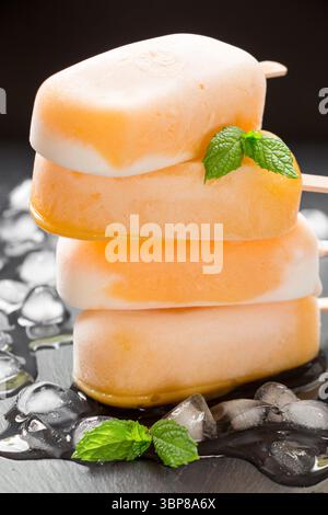 Three mango ice cream popsicles are neatly stacked on top of each other, resting on crushed ice. Fresh mint leaves garnish the popsicles, enhancing the vibrant colors and inviting a fresh summer vibe. Stock Photo