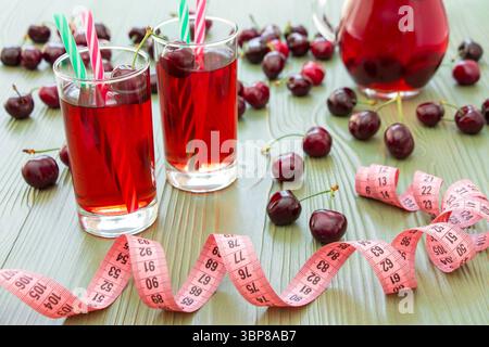 Left 2 glasses of juice with straws, scattered cherries, jar of cherry juice, in front centimeter on light green background. Cherries, cherry juice and centimeter. Horizontal. Stock Photo
