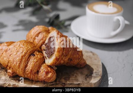 Food, Fresh chocolate croissants on a rustic wooden board beside a cup of cappuccino, with a soft grey background Stock Photo