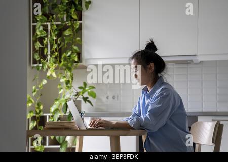 Focused Asian woman freelancer working remotely sits at table with laptop in kitchen own home. Self-sufficient young Korean female copywriter is typin Stock Photo