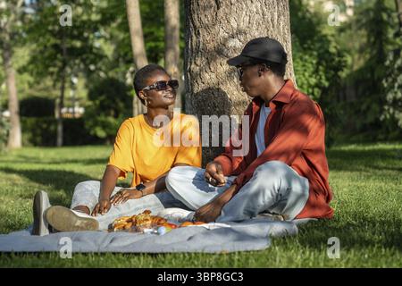 Contented african american loving couple rest on grass looking for each other, chatting have picnic good sunny weather together. Happy relaxed black g Stock Photo