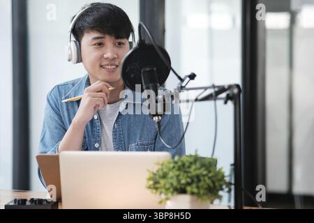 A man wearing headphones and a blue shirt is sitting at a desk with a laptop and a plant. He is smiling and he is focused on his work Stock Photo