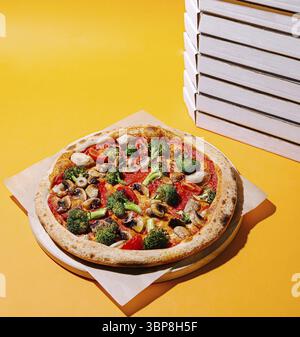 Food, A delicious veggie pizza topped with colorful ingredients sits on a wooden board. Stacks of empty pizza boxes in white are neatly arranged nearb Stock Photo