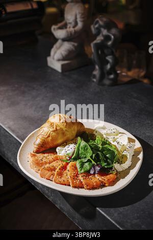 Food, A plate features thinly sliced smoked salmon alongside fresh greens and a croissant, all artistically arranged on a dark countertop creating a w Stock Photo