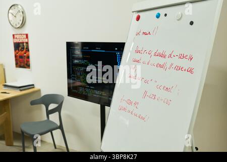 Whiteboard displaying handwritten code standing in front of monitor showing programming interface in modern classroom setting with empty chairs and educational poster on wall Stock Photo