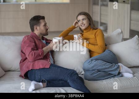 Happy joyful woman sitting with interested pleased man husband on home couch enjoy coffee, chatting planning future. Married loving couple leisurely t Stock Photo