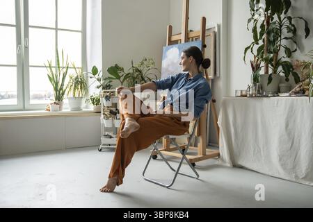 Expressive relaxed woman painter sits cross leg looks out window in art workshop with paintings takes break from creative work. Thoughtful girl artist Stock Photo