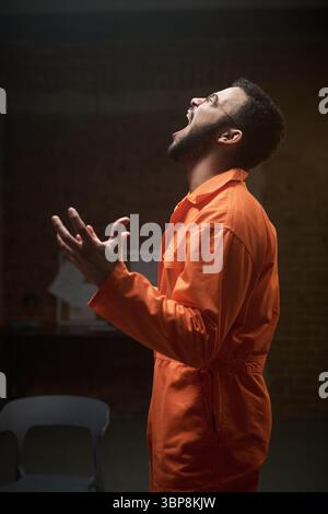 Young adult man wearing prison uniform standing in group therapy room shouting with hands raised, expressing intense emotion during rehabilitation session in correctional facility Stock Photo