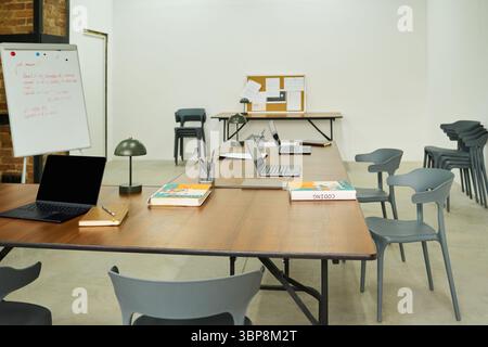 Empty classroom with several laptops, notebooks, and pens arranged on long tables, chairs surrounding workspace, whiteboard and bulletin board visible in background, educational setting in prison Stock Photo