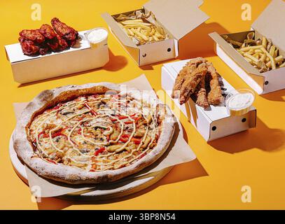 Food, A large pizza topped with red peppers and mushrooms sits alongside crispy chicken wings and golden fries, all arranged in takeout boxes on a vib Stock Photo