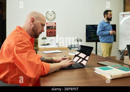 Caucasian man with shaved head and tattoos working on laptop at desk, Caucasian young adult man with beard standing near whiteboard presenting coding information Stock Photo