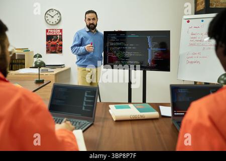 Middle aged Caucasian man explaining computer programming code on large monitor to group of young adults wearing orange uniforms using laptops in classroom Stock Photo