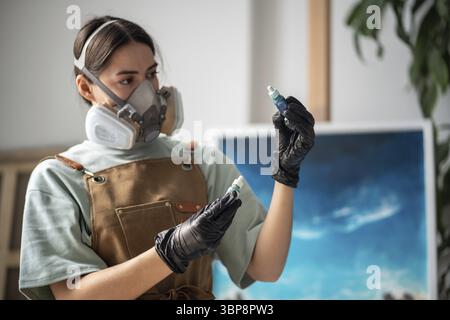 Attentive female artist choosing pigments to work at epoxy resin painting. Girl in respirator making choice between two blue paints, picking most suit Stock Photo