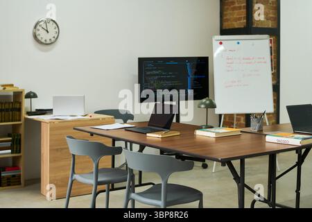 Empty classroom with laptops, textbooks, digital screen displaying code, whiteboard with handwritten notes, clock on wall, setting representing prison education environment Stock Photo