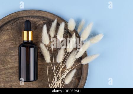 Blank amber glass essential oil bottle with pipette on round wooden tray on blue background decorated lagurus bouquet. Skin care concept with natural Stock Photo