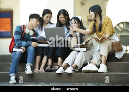 A group of college students studying together outdoors on campus, using a laptop and books in a collaborative setting Stock Photo