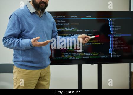 Middle aged Caucasian man teaching programming in prison classroom, holding marker and gesturing in front of large screen displaying code, focusing on educational instruction Stock Photo