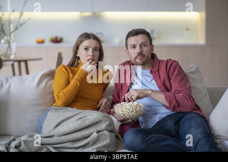 Amazed couple sitting on couch with popcorn, watching TV program, favourite show, film attentively, shocked, surprised by unexpected episode. Wife and Stock Photo