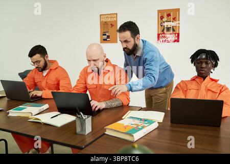 Middle aged Caucasian man instructing group of adult men working on laptops in classroom setting, participating in educational program Stock Photo