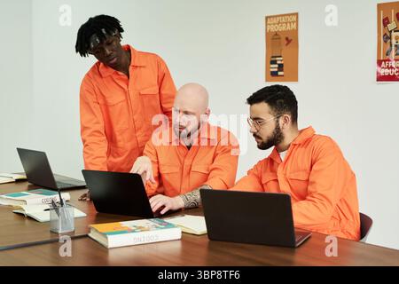 Three young adult men collaborating at table using laptops and books, discussing project in classroom setting Stock Photo