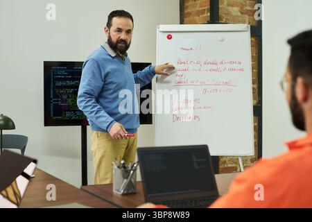 Middle aged Caucasian man teaching programming concepts on whiteboard to adult male student in classroom setting, discussing code structure with laptops and digital screen visible Stock Photo