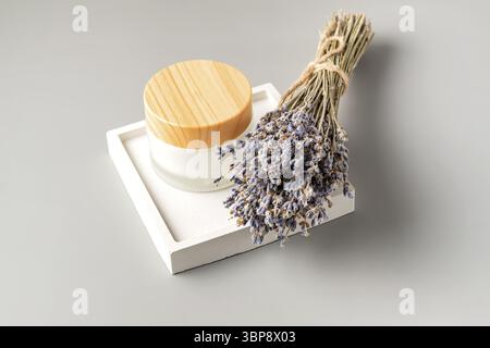 White frosted glass jar with cream of lavandula, flowers bouquet of dried purple lavender blossom on concrete square tray. Herbal aromatherapy and ski Stock Photo