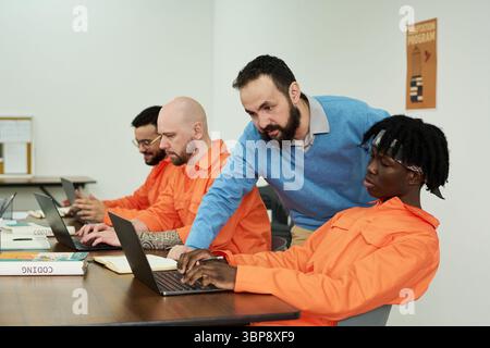 Middle aged Caucasian man assisting group of young adult men working on laptops in classroom setting, focusing on coding tasks Stock Photo