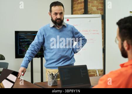 Middle aged Caucasian man standing and instructing young adult man with beard in orange prison uniform, during educational session in classroom with whiteboard and computer monitors Stock Photo