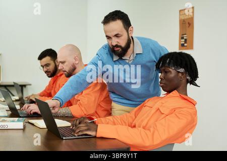 Middle aged Caucasian man instructing group of young adult and middle aged men of diverse ethnicities working on laptops in classroom setting, teacher guiding student at desk Stock Photo