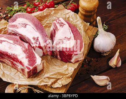 Food, Three cuts of fresh meat rest on crumpled paper atop a wooden board surrounded by whole garlic bulbs and ripe cherry tomatoes, creating a rustic Stock Photo