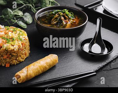 Food, Delectable meal of fried rice, spring roll, and spicy soup served on a sleek black slate Stock Photo