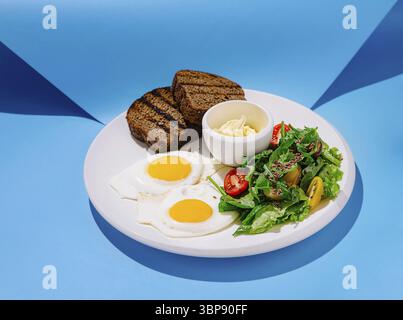 Food, A vibrant breakfast plate features two sunny-side-up eggs, a fresh salad with tomatoes, and slices of toasted bread, all served against a blue b Stock Photo