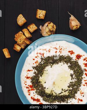 Food, Turquoise plate with creamy yogurt dip decorated with herbs and spices, served with toasted bread croutons and garlic on dark wooden background Stock Photo