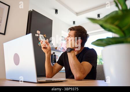 Man working on laptop at home and using remote control to adjust temperature on air conditioner. Person struggling with heat while working at home off Stock Photo