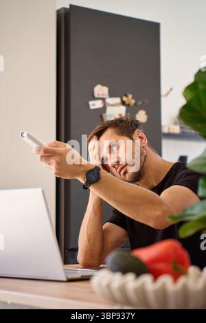 Man working on laptop at home and using remote control to adjust temperature on air conditioner. Person struggling with heat while working at home off Stock Photo