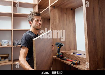 Man assembling wooden wardrobe at home using tools and holding large wooden panel. Concept of DIY furniture assembly, home improvement and renovation Stock Photo