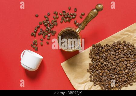 Top view of fresh coffe prepared in cezve (traditional turkish coffee pot), whte cup and coffee beans on craft paper on red background Stock Photo
